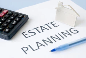 The image shows the words "Estate Planning," and around those words you see a ballpoint pen, a miniature house, and an electronic calculator.